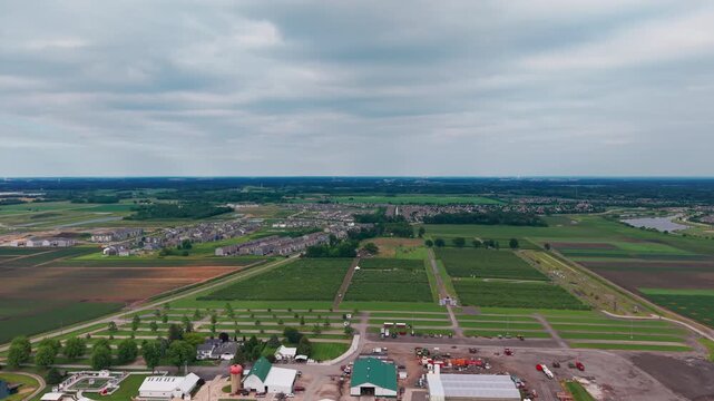 Aerial drone view of a suburban neighborhood surrounded by farmland and open agricultural fields under an overcast sky. Wide panoramic landscape showcasing residential development, rural countryside.