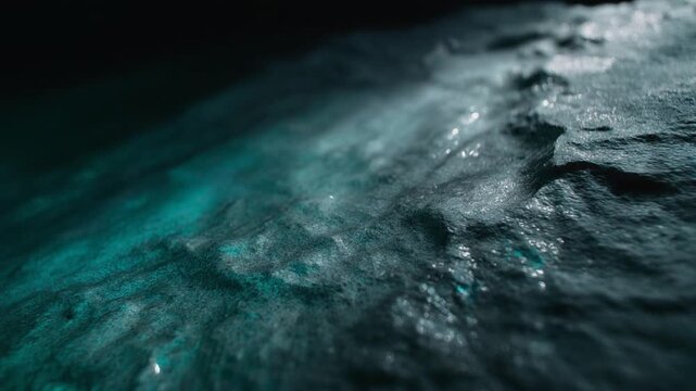 Colored water slowly washes across textured wet stone, forming thin reflective sheets through deep grooves under dramatic angled light in a moody teal and slate macro scene.