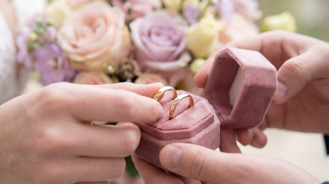 Couple exchanging gold wedding rings from pink velvet box near pastel bouquet, marriage ceremony, commitment, romance, bridal detail, suitable for editorial themes, marketing campaigns,