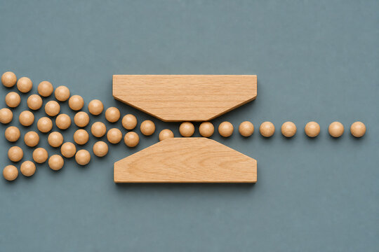 Wooden beads move through narrowing hourglass pathway, illustrating business bottleneck, workflow constraints, process delays, operational inefficiency and restricted productivity in minimalist flat l