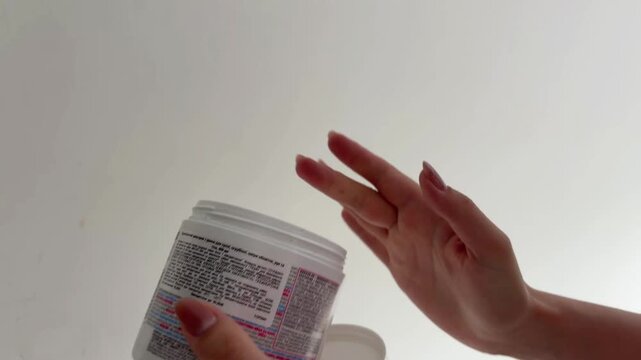 Woman with manicured nails opening a cosmetic jar and scooping out a white face cream for her skincare routine, showing the texture of the product isolated on a white background