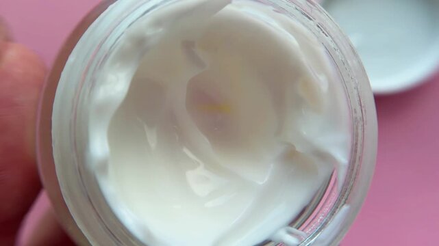 Woman's hand holding a clear glass jar of luxurious white face cream, displaying its smooth and rich texture, with the white lid resting on a vibrant pink background
