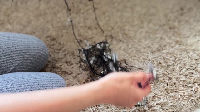 Woman's hands carefully untangling knotted black wire of christmas fairy lights, preparing festive home decorations on a soft beige carpet before the holiday season