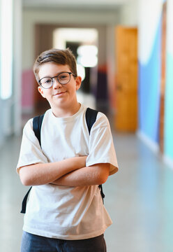 Smiling pupil wearing eyeglasses and backpack crossing arms in school hallway