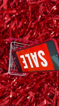 Miniature shopping cart with a smartphone displaying sale text moving over a red tinsel background, symbolizing online shopping discounts, black friday promotions, and e commerce