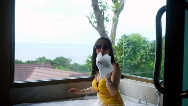 Asian woman in yellow swimsuit and sunglasses enjoying a relaxing bubble bath in a luxury resort ; large glass window overlooks a scenic sea view and lush green trees