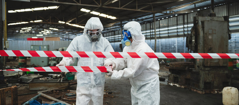 Two workers in full protective gear stand behind a red-and-white barrier
