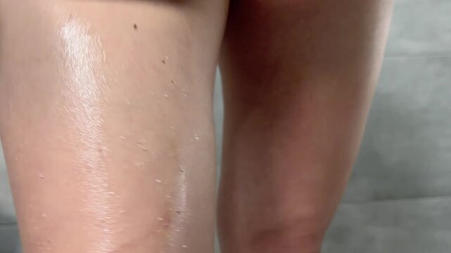 Woman massaging her legs with anti cellulite scrub for skin care, health, and beauty. Young female person applying exfoliating cosmetic product on thighs in the bathroom