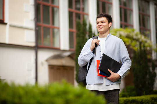 Student stands outside school building holding books and laptop while looking around on a sunny day during spring season