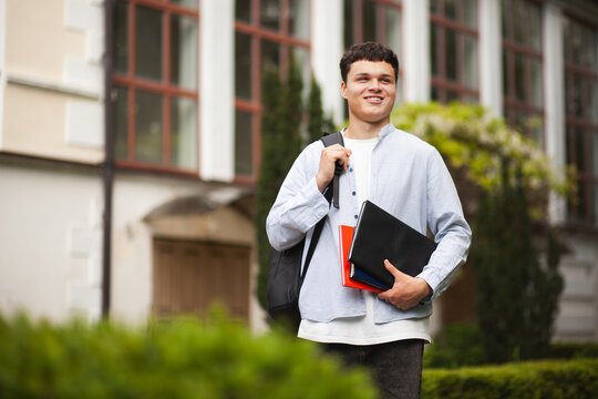 Young student stands outside a building with books and a bag on a sunny day