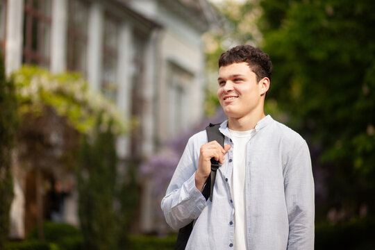Young man stands outside a building with trees during daytime while holding a backpack strap