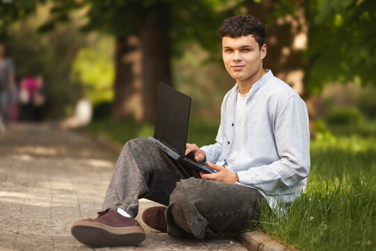 Young man sits on path in park using laptop while surrounded by trees during daytime