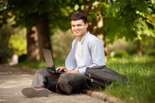 Young man sits on pathway using laptop under trees in a public park during daytime