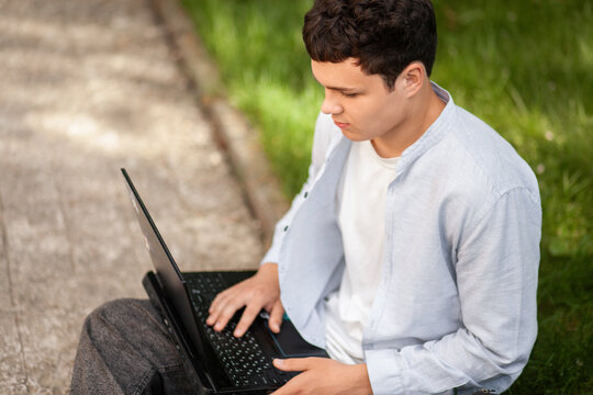 Young man works on laptop outside in green park during daytime while sitting on ground