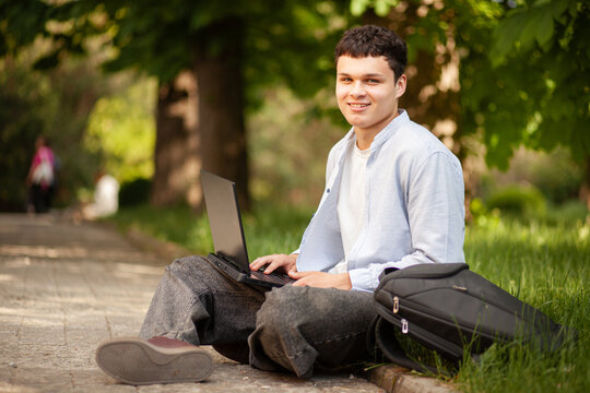Young man works on laptop outdoors in park during sunny day while sitting on path with backpack nearby