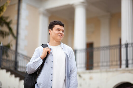 Young man walks near building wearing backpack and smiling while looking forward on sunny day in city square