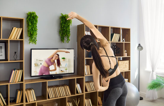 Woman stretching at home with online workout. She follows a fitness class on TV, bending to boost flexibility in a bright room with shelves and plants. Concept of home fitness lifestyle.