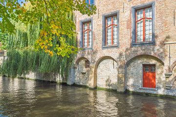 Obraz premium Bruges town in Belgium, Europe. Historic brick building with arched foundations along a serene canal, lush green foliage cascades over the water, under warm sunlight