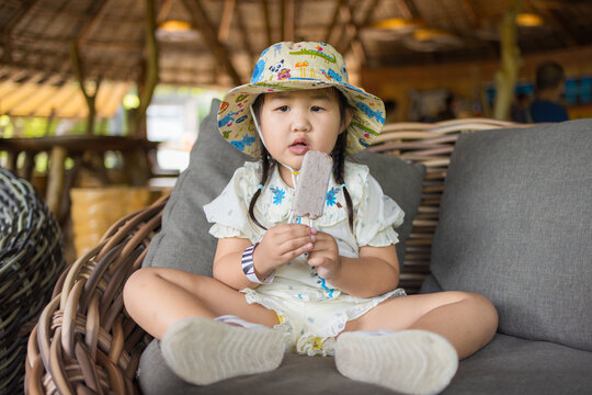 Cute Little Girl Eating Ice Cream on a Sofa,A cute little Asian girl wearing a patterned bucket hat sitting comfortably on a rattan sofa and enjoying a delicious ice cream pop.