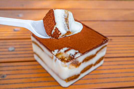 Spoon lifting slice of tiramisu dessert with cream and cocoa layers on wooden table, Italian food concept