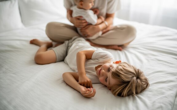 Young mother holding her newborn baby while lying on a comfortable white bed with her young son resting nearby, capturing a peaceful and loving family morning moment