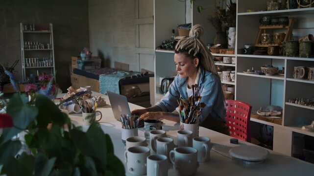 Ceramic shop owner types on laptop at desk with handmade stuff