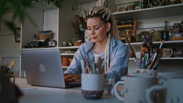 Craftswoman works on laptop against shelves with ceramic stuff