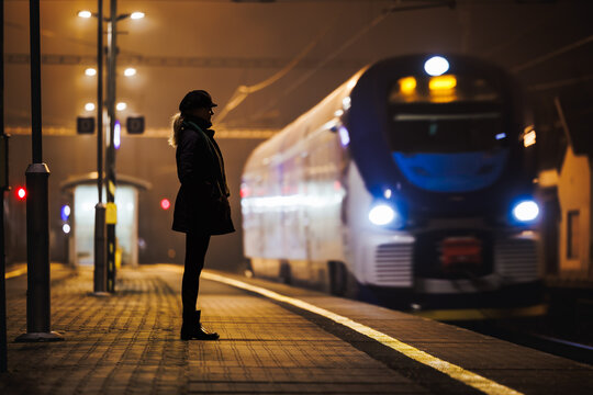 Woman looking at arriving train at empty railway station at night. City life and commute by public transportation