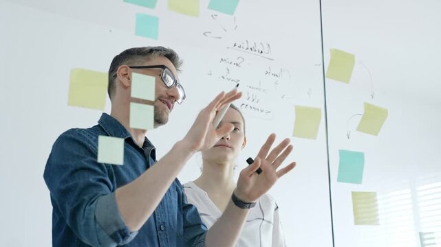 Colleagues collaborating on a clear glass board, brainstorming business strategy, developing innovative ideas, and organizing project tasks with colorful sticky notes in a modern office
