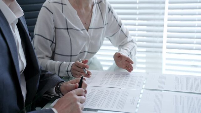 Business professionals reviewing a contract document at a table. Pointing at clauses with a pen. Discussing legal terms and financial agreements during an official meeting