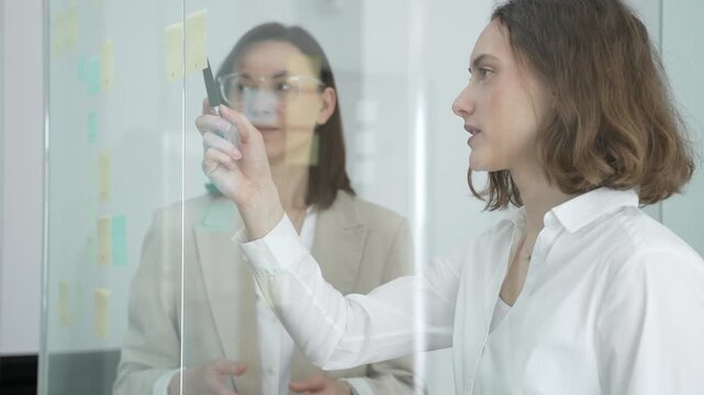 Analyzing project strategy and collaborating on new ideas, businesswomen brainstorming with sticky notes on a glass board during an office meeting, planning future development