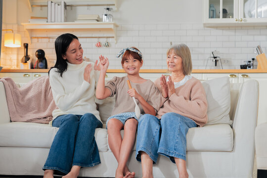 A little girl sings into a wooden spoon microphone while her mother dances and grandmother claps along on the sofa a playful, joyful family moment of impromptu music and shared laughter at home.