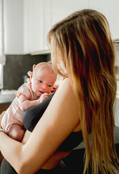 Mother gently cradles her small newborn baby in soft morning light within home kitchen setting