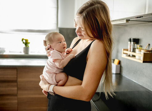Young woman holds newborn child close inside kitchen during bright morning daylight hours