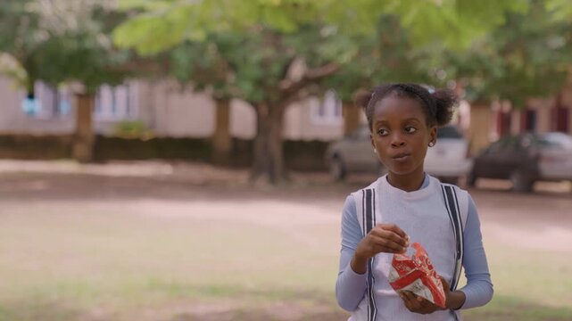 Black daughter eating snack near playground schoolage girl with backpack holds chip bag, expressive look while chewing, mother in red approaches from side, campus trees and parked cars in background,