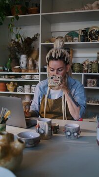 Female potter enters shop data at laptop and drinks tea in studio