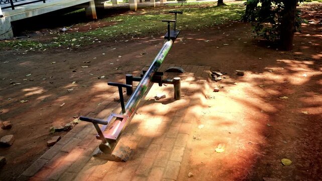 An empty metal seesaw stands in a quiet public park playground on a sunny day, casting shadows on the ground with fallen leaves.
