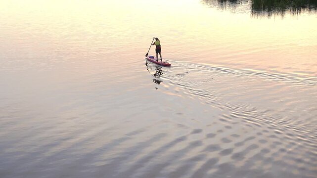 High angle footage shows a lone paddleboarder standing and paddling across calm reflective water at sunset, leaving gentle ripples and a soft mirrored silhouette