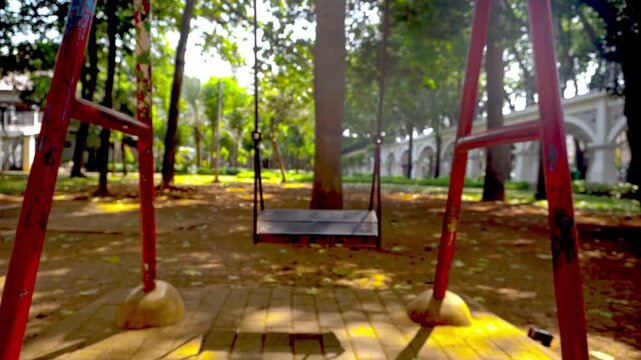 An empty metal playground swing moves gently on its own in a quiet public park on a bright sunny day.