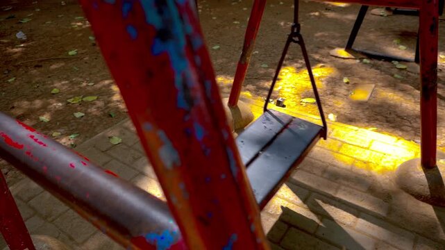 An empty metal playground swing moves gently on its own in a quiet public park on a bright sunny day.
