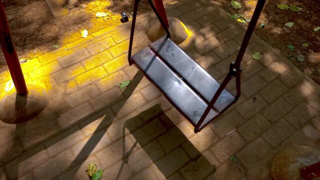 An empty metal playground swing moves gently on its own in a quiet public park on a bright sunny day.