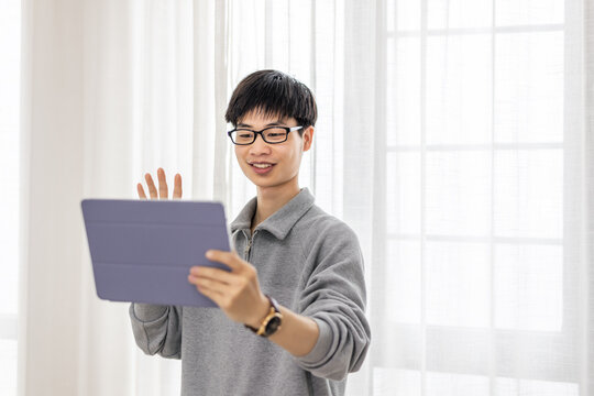 A young asian man with glasses waves while holding a tablet for a video call in an indoor space.