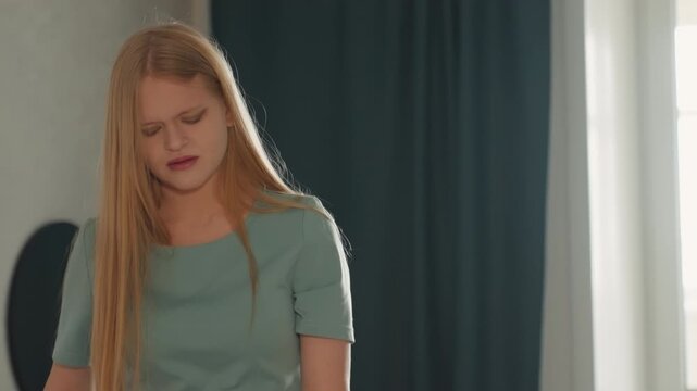 Frustrated white woman during house cleaning, standing in living room near window and dark curtain, long blonde hair, teal top, pained expression, slumped shoulders, visible fatigue, domestic chores