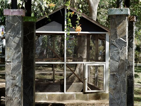 Park birdhouse cage framed by stone pillars with hanging orange flowers and green trees in daylight.