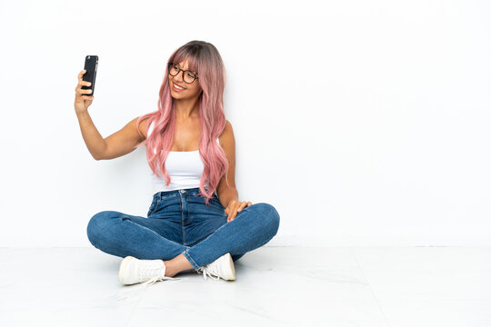 Young mixed race woman with pink hair sitting on the floor isolated on white background making a selfie