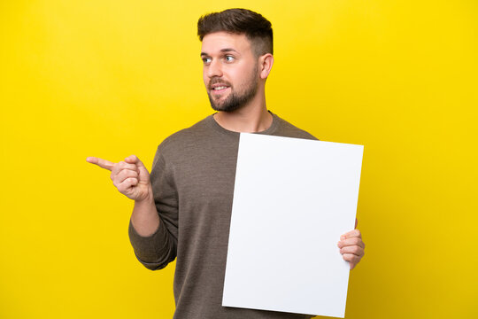 Young caucasian man isolated on yellow background holding an empty placard and pointing side