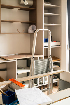 Setting up wooden cabinetry at a home improvement site using a step ladder for reach. Workspace features multiple shelves, tools, and building materials visible. New kitchen concept. 