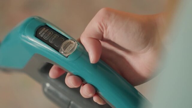 Hand presses vacuum mode dial during house cleaning, professional cleaner tests power and mode on teal handheld vacuum with steady motion, closeup on button and display shows setting change