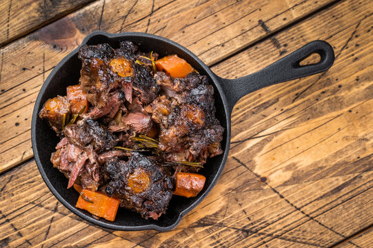 Braised beef oxtail stew with carrots and rosemary in cast iron skillet on rustic wooden table, rich dark sauce
