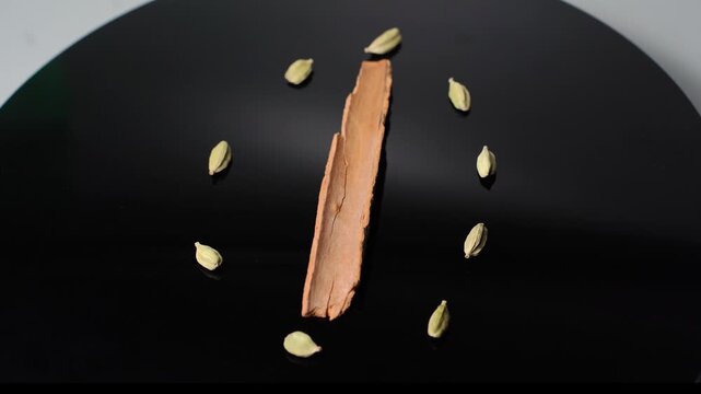 Time concept: Cinnamon Stick and Cardamom Pods Rotate on a Black Dish, Evoking Culinary Spices, Cooking, and Aromatic Ingredients for Delicious Dishes